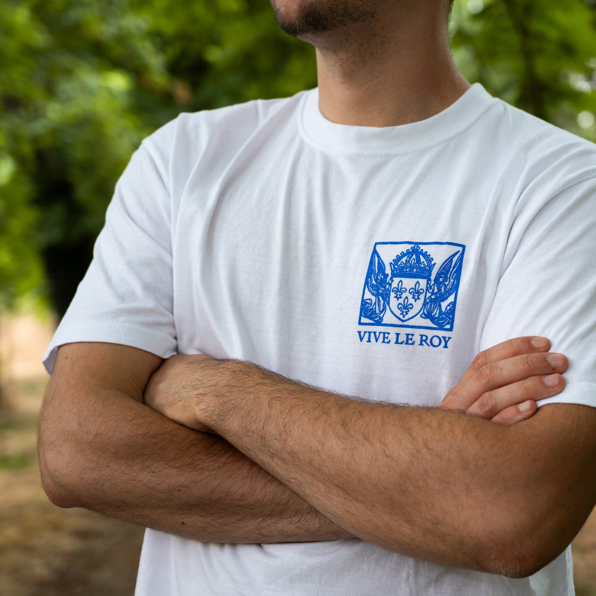 Personne portant un t-shirt blanc « Vive Le Roy », les bras croisés, debout à l'extérieur.