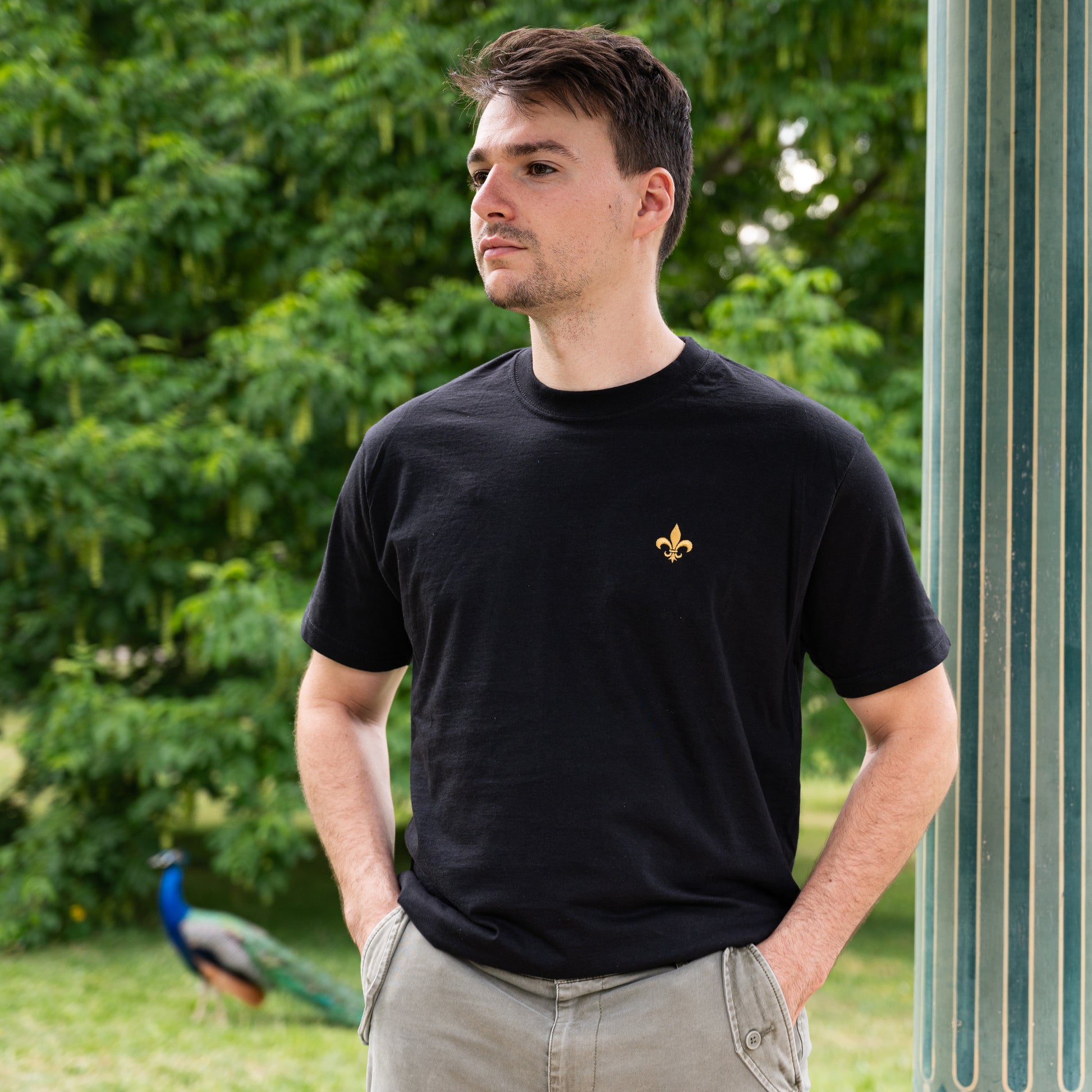 Homme portant un t-shirt unisexe noir avec emblème doré, debout à l'extérieur.